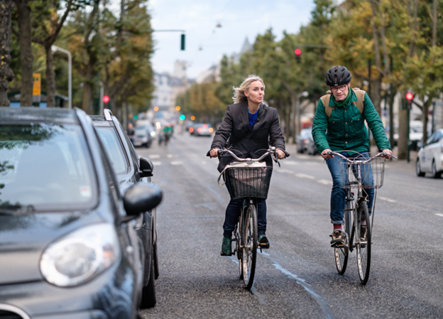 Two commuters, a middle-aged woman and man, on bicycles on a wide street.
