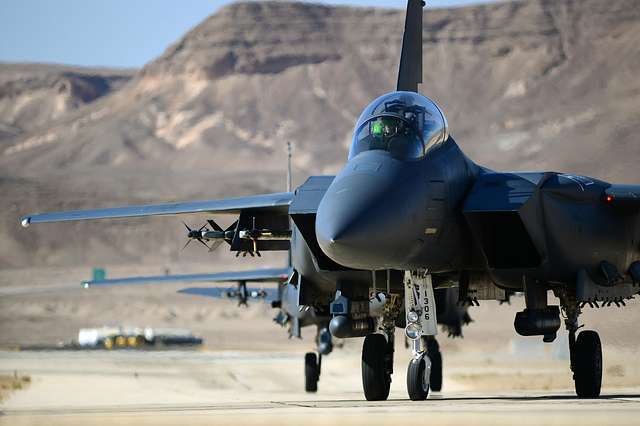 Black fighter jet on a runway, with a backdrop of desertous mountains.