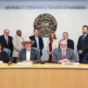 council-members-chambers Four people sit at a long desk with six people standing behind them in front of the county crest.