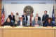 Four people sit at a long desk with six people standing behind them in front of the county crest.