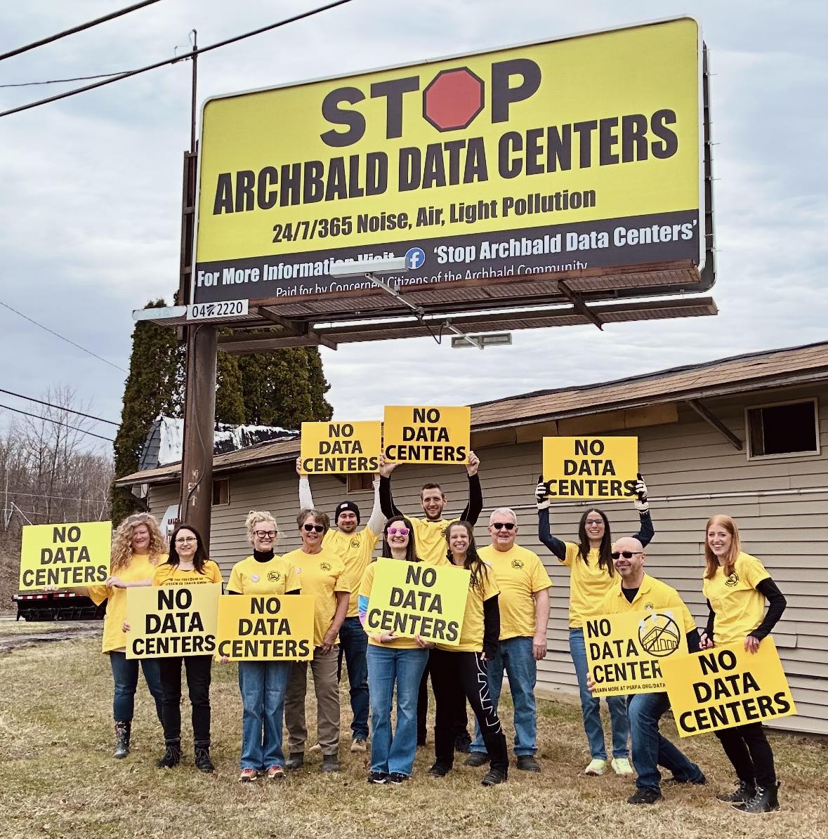 a group of people stand under a billboard, smiling and holding signs