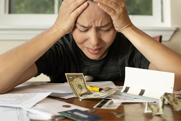 Woman looking stressed at a pile of papers and dollar bills, holding her head in her hands.