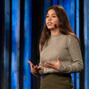 A young, professionally dressed person speaks on a stage.