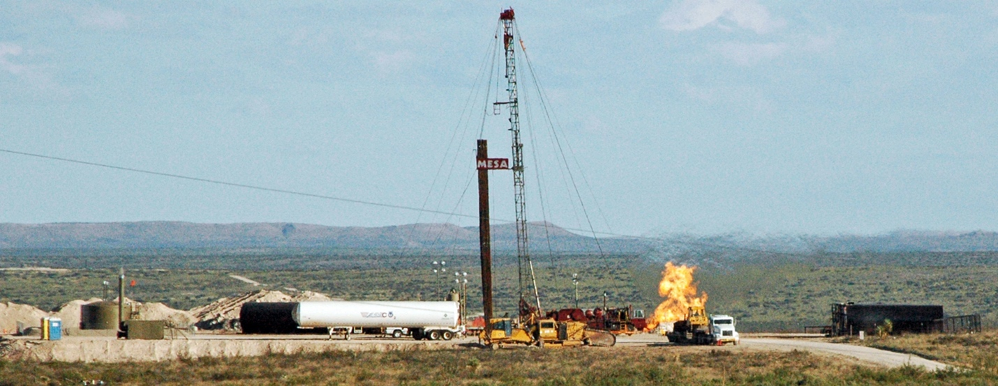 A drilling site with infrastructure, vehicles, and a large fire in the middle of wide-open plains with mountains in the distance.