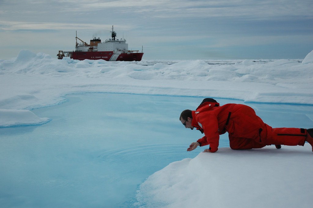 image of a researcher exploring an Arctic pool, with an icebreaker ship in the background, cutting through Arctic ice.