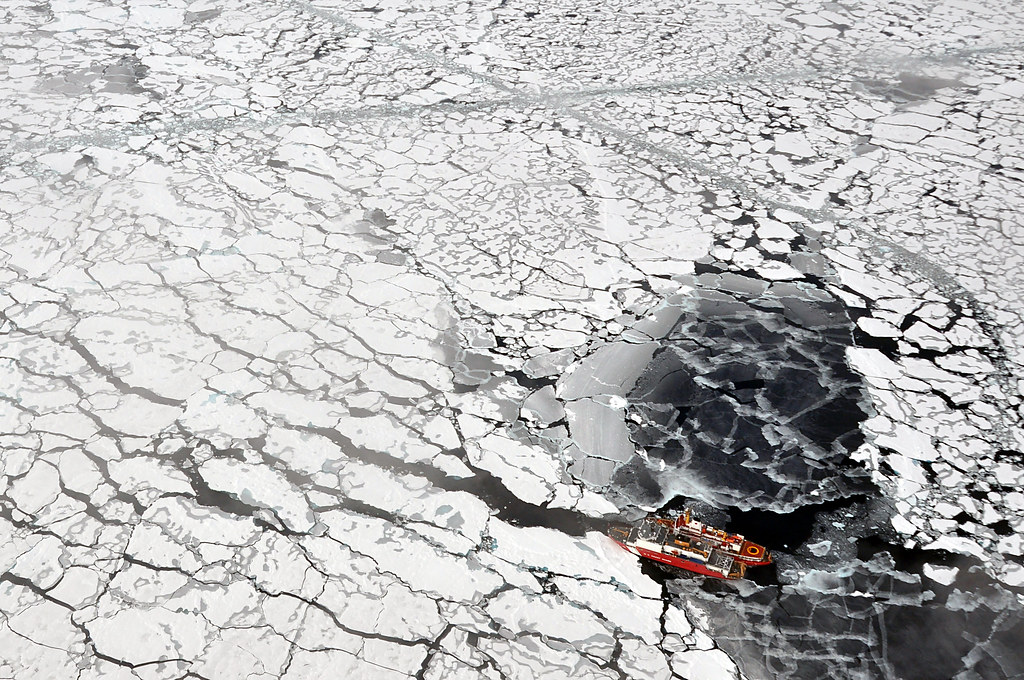 Birds-eye shot of an icebreaker ship in the Arctic, with patches of cracked ice floating atop the sea.