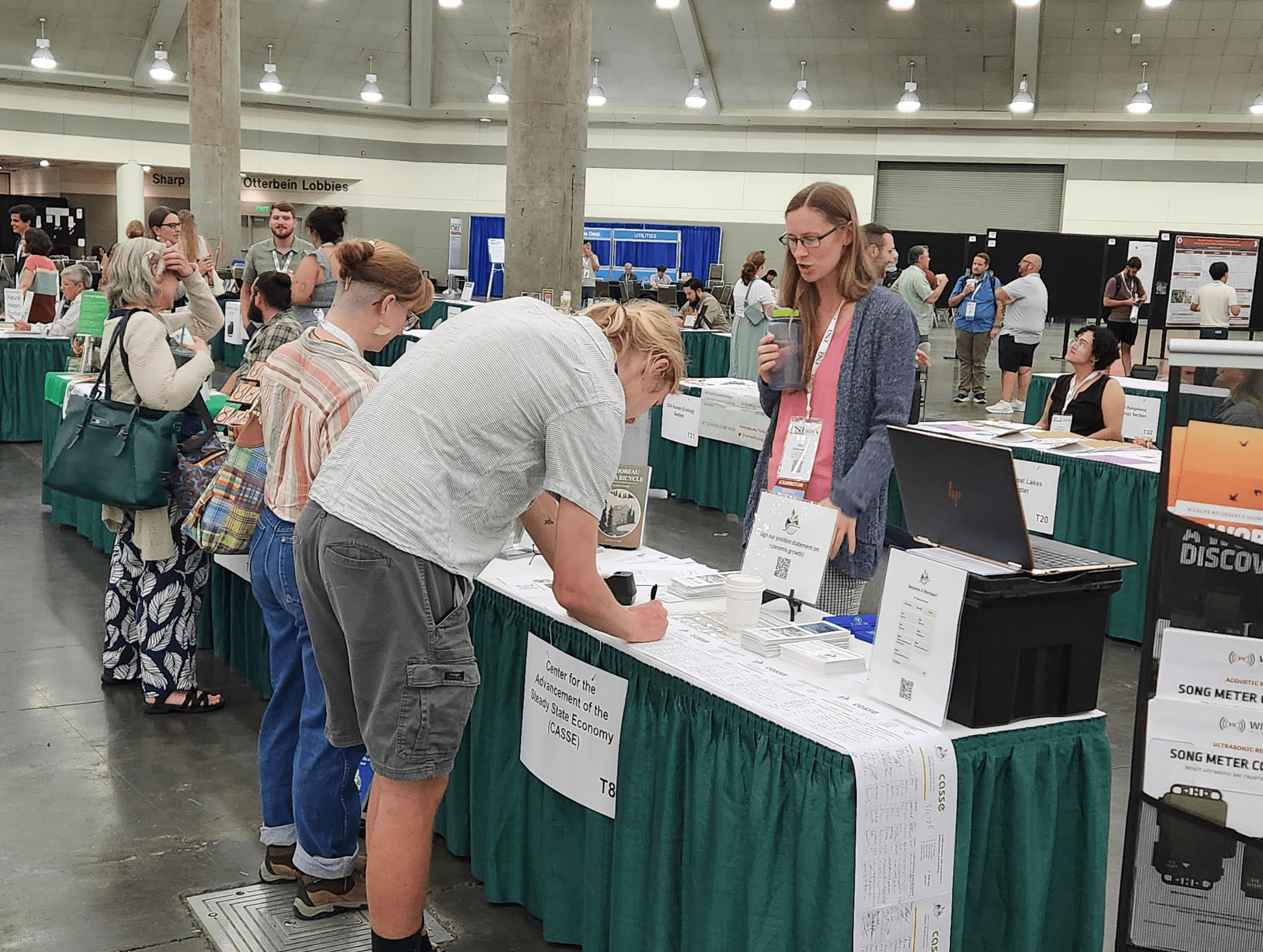 Three people stand at a table in a conference hall. The person behind the table is talking to the other two, and one person leans over to sign a paper.