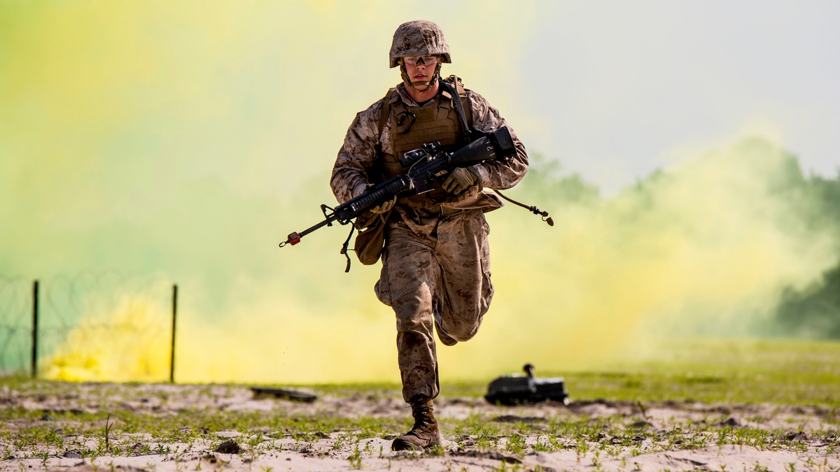 U.S. soldier carrying a big gun and running toward the camera, with yellow smoke in the background.