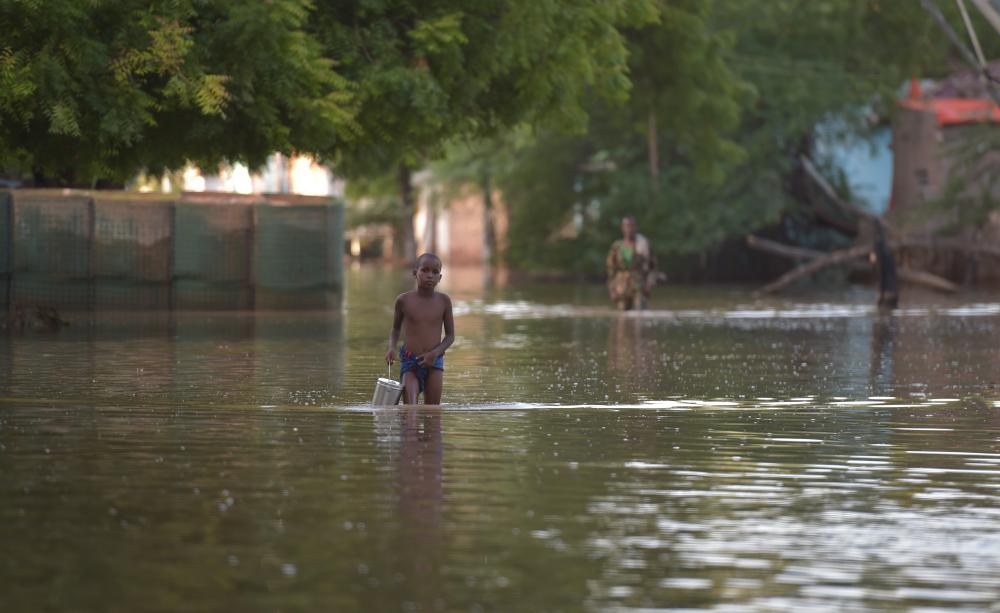 A young boy wades through thigh-deep water, carrying a bucket. A woman also wades through in the background. There are partially submerged buildings around.