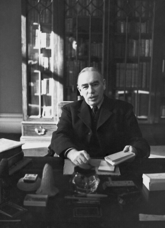 black-and-white photo of Keynes at a desk, holding a notebook, with a bookshelf behind him