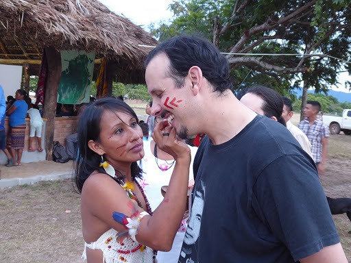 A man (this article's author) smiles as an Indigenous woman paints his face, with a thatched hut in the background.