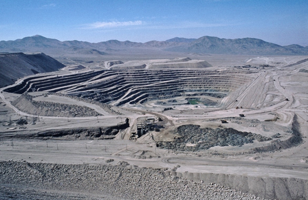 Aerial view of an open-pit mine in an arid region, with mountains in the background.
