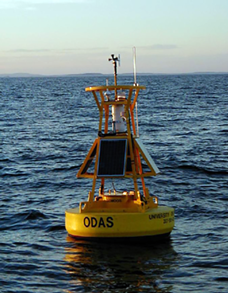 A yellow buoy equipped with solar panels and measurement instruments sits on a vast body of water with mountains in the far distance.