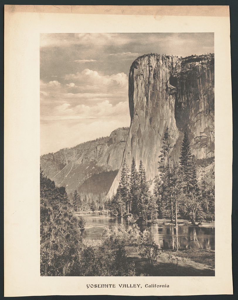 An old sepia image of a huge cliff face with a body of water at its feet.