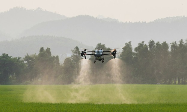 A drone sprays liquid onto an even, green field, with trees and mountains in the background.