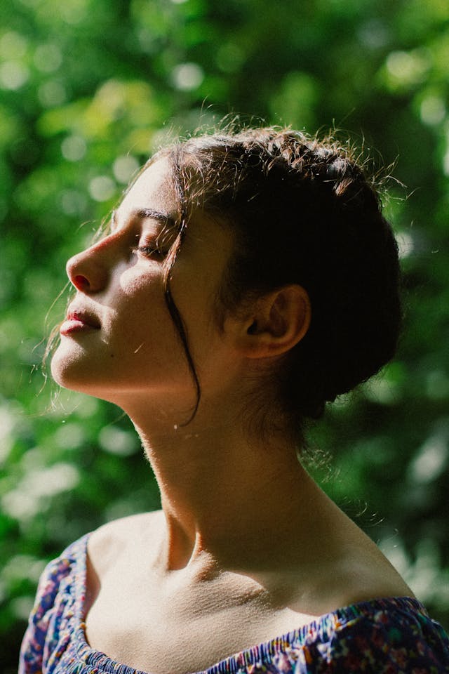 A young person closes their eyes as sunlight hits their face, with green leaves in the background.