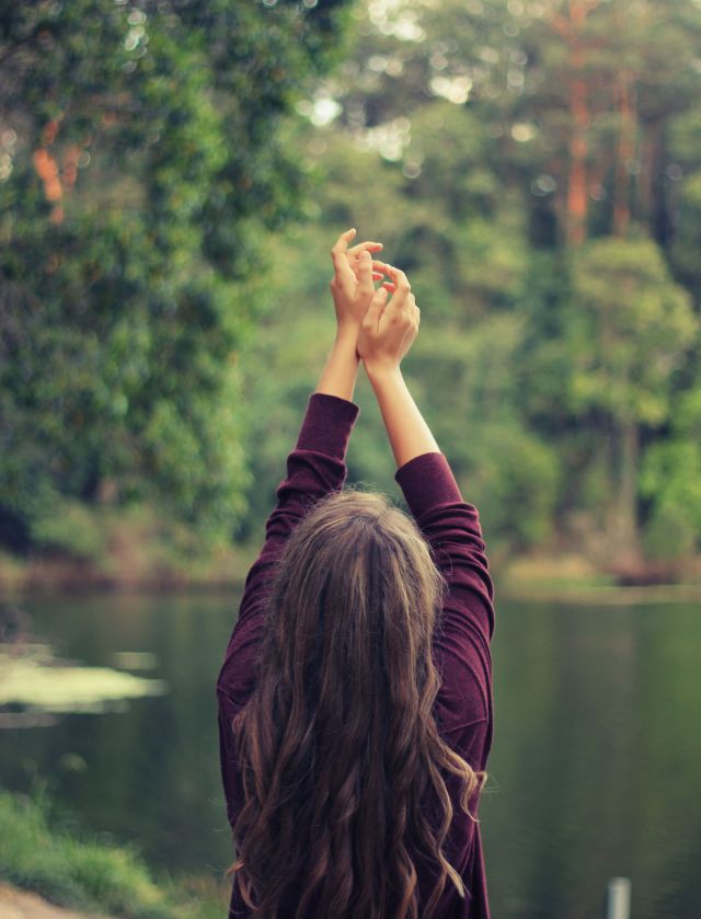 the back of a young person with her arms raised, surrounded by nature