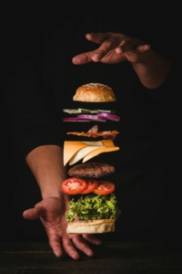 Image of a burger spread out vertically, with space between layers of bun, onions, cheese, the burger, tomatoes, and lettuce. A man's hand, palm down, is above the top bun, and his other hand, palm up, is under the bottom bun. The layers appear to be floating in the air.