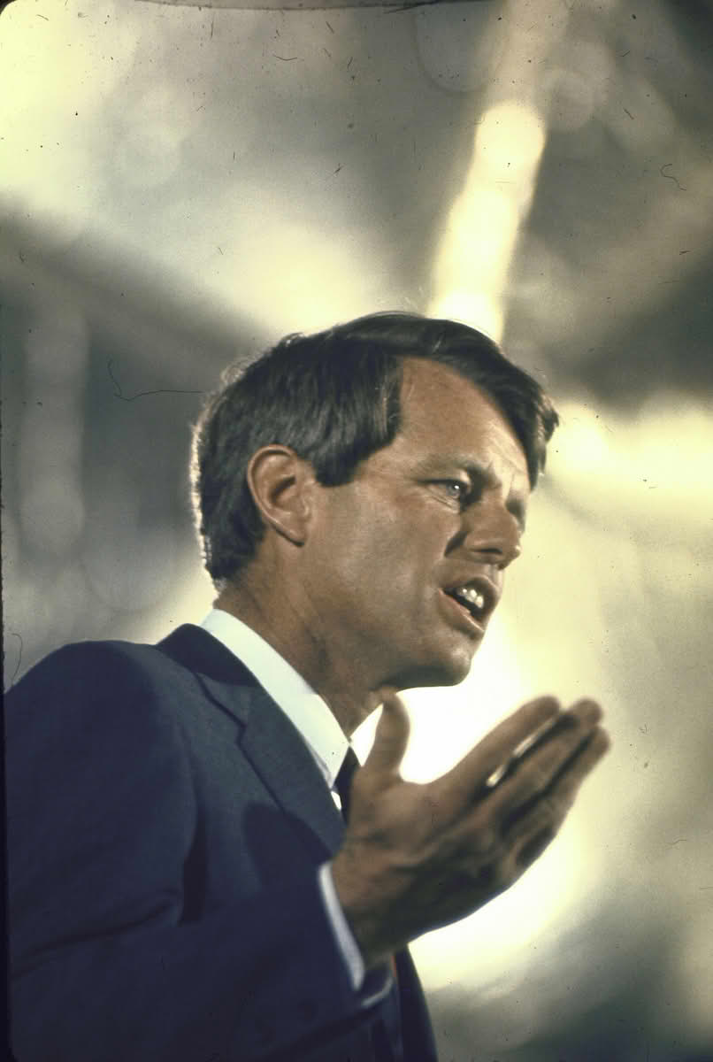 Kennedy speaks under a spotlight with a hand partially raised