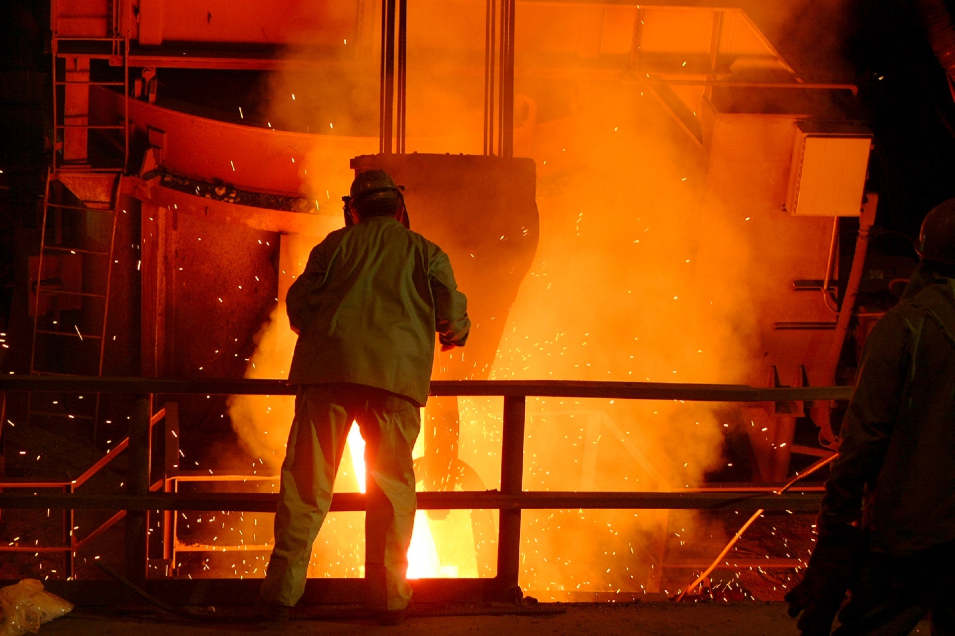 A worker with protective gear leans over a railing, with big machinery and white-hot fire in front of them.