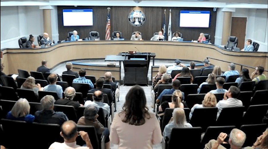 Someone stands in the foreground in an aisle facing supervisors seated at the front of the room (background of the image). People sit in chairs on either side of the aisle, also facing the supervisors.
