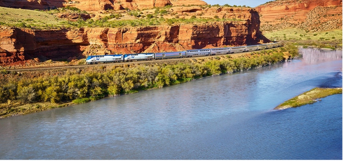 long passenger train rounds a bend with red cliffs in the background and a river in the foreground