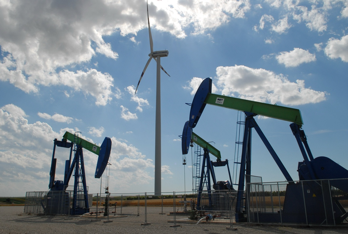 Three oil pumpjacks in the foreground and a windmill in the background towering over them.