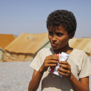 A thin young boy stands in the foreground, eating food from a plastic wrapper, with gravel and tan tents in the background.
