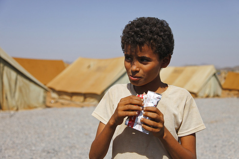 A thin young boy stands in the foreground, eating food from a plastic wrapper, with gravel and tan tents in the background.